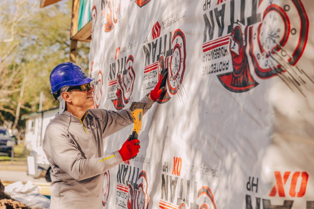 Man in a hardhat, volunteer, uses a stapler to attach wrap to a house